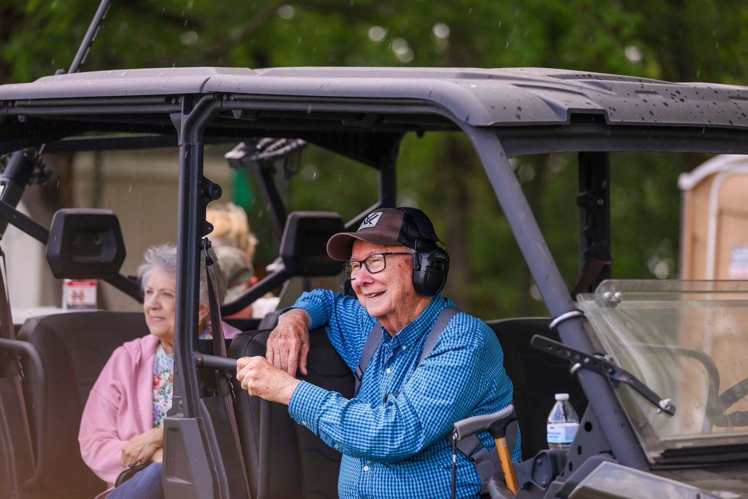 Smiling senior man in blue jacket and cap leans on UTV vehicle outdoors, with woman in pink visible behind him.