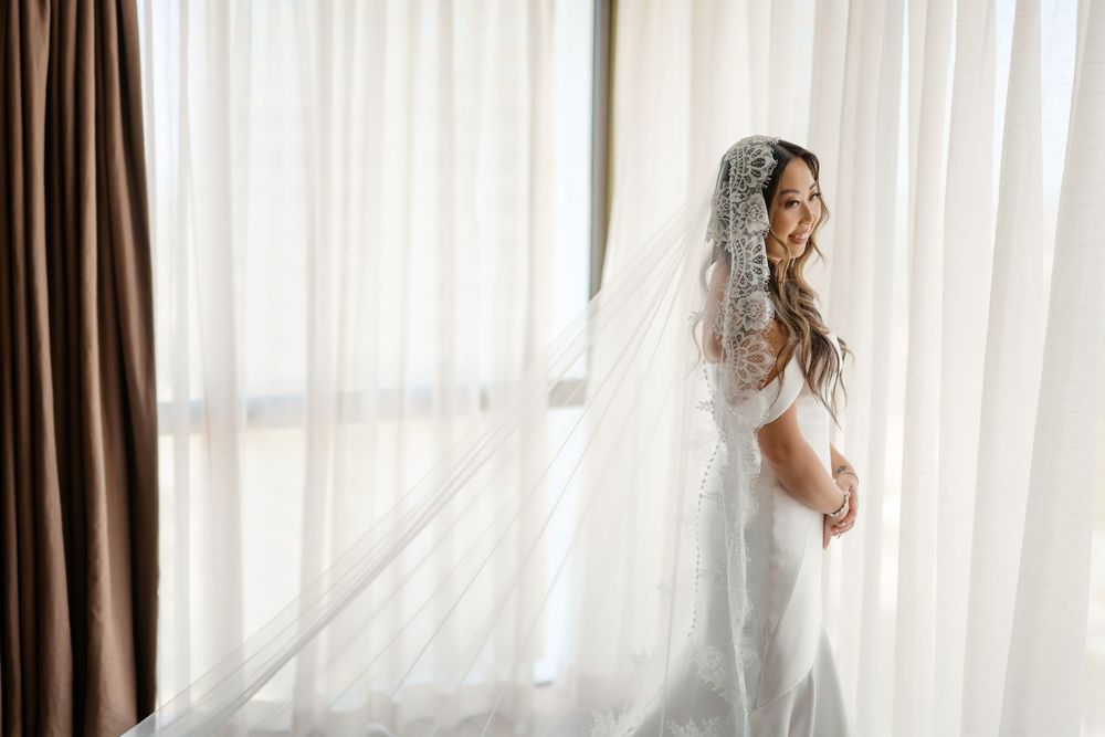 Smiling bride in white lace wedding dress and cathedral veil standing by sheer curtained window in soft natural light.