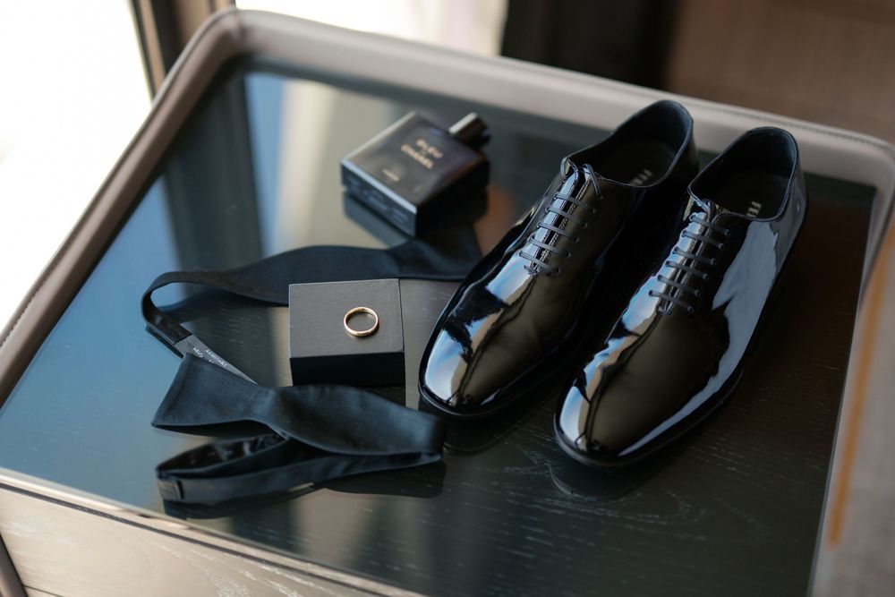 Black patent leather dress shoes, bow tie, cufflinks, and ring displayed on glass surface for a formal wedding.