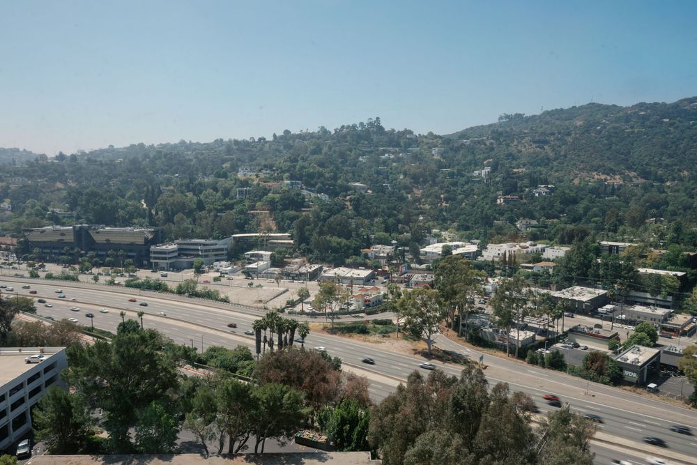 Aerial view of a Los Angeles freeway surrounded by lush green hills, trees, and urban buildings on a clear day.