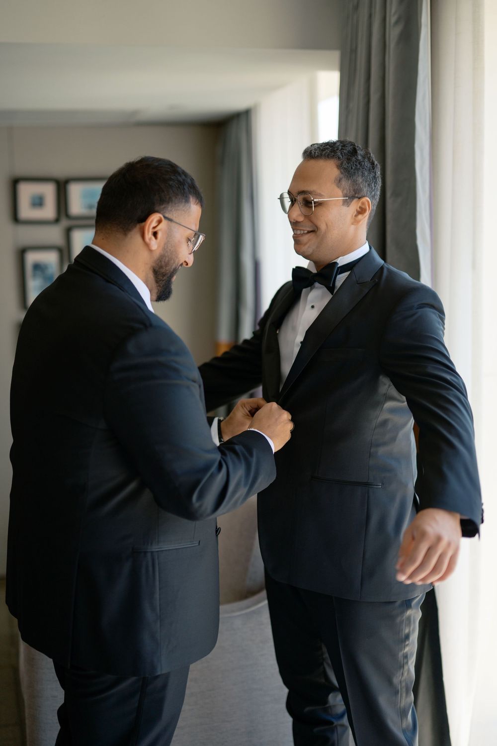 Two men in black tuxedos help each other get ready for a wedding ceremony indoors.