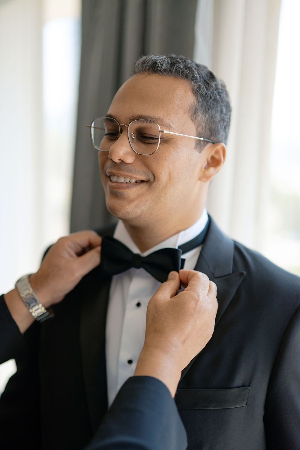 Smiling groom in black tuxedo and glasses has his bow tie adjusted before wedding.
