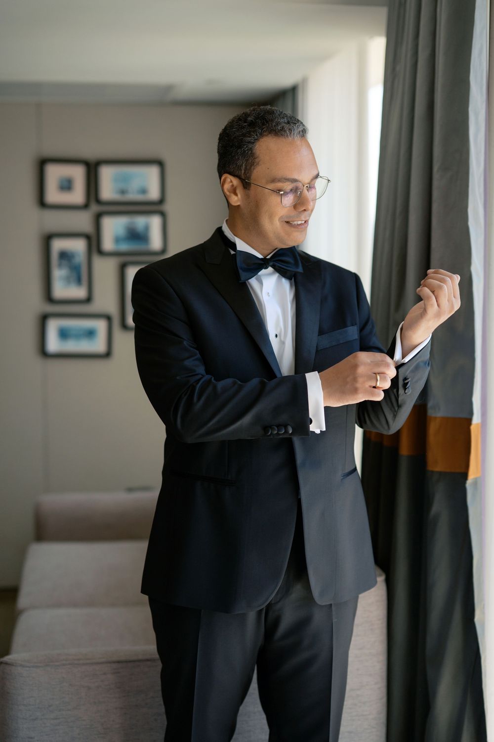 Groom in black tuxedo and bow tie adjusting cufflinks by window before wedding ceremony.