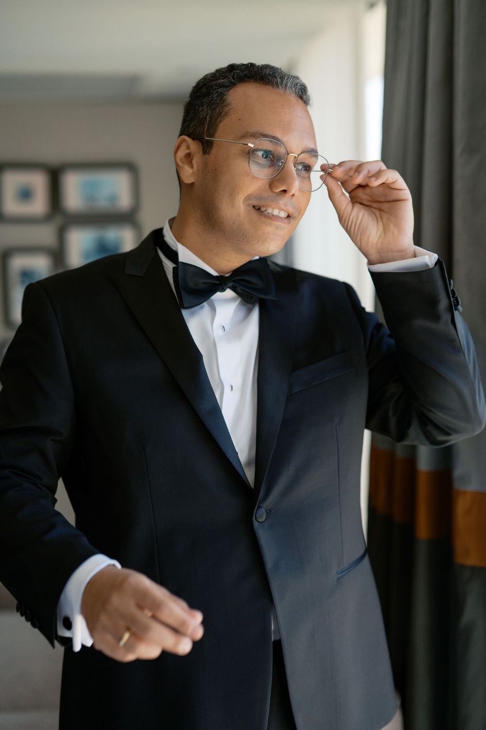 Dapper groom in black tuxedo and bow tie adjusting his glasses while smiling indoors.