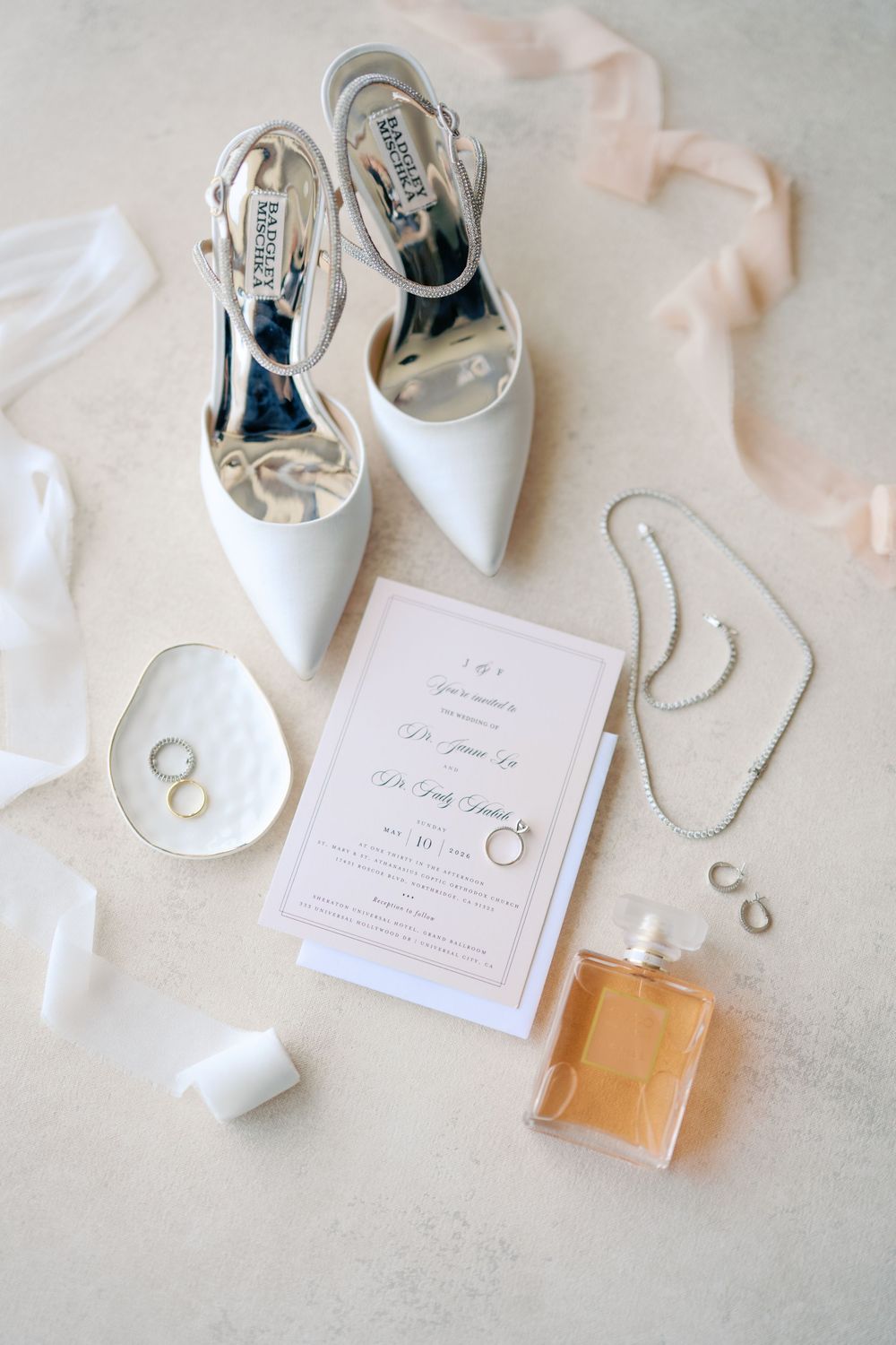 Elegant bridal flat lay featuring white heels, wedding invitation, perfume, jewelry, and rings on a neutral background.