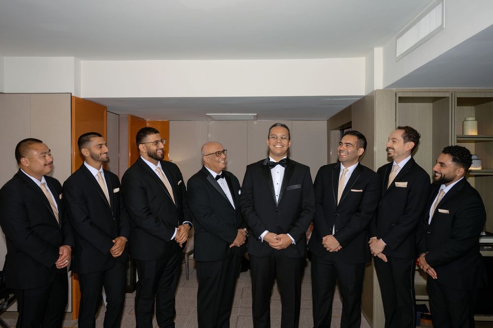 Groom and groomsmen in black tuxedos laughing together before wedding ceremony.