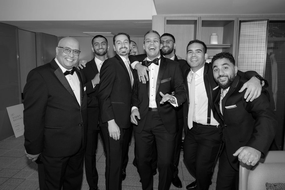 Groom and groomsmen in black tuxedos celebrate together before wedding reception.