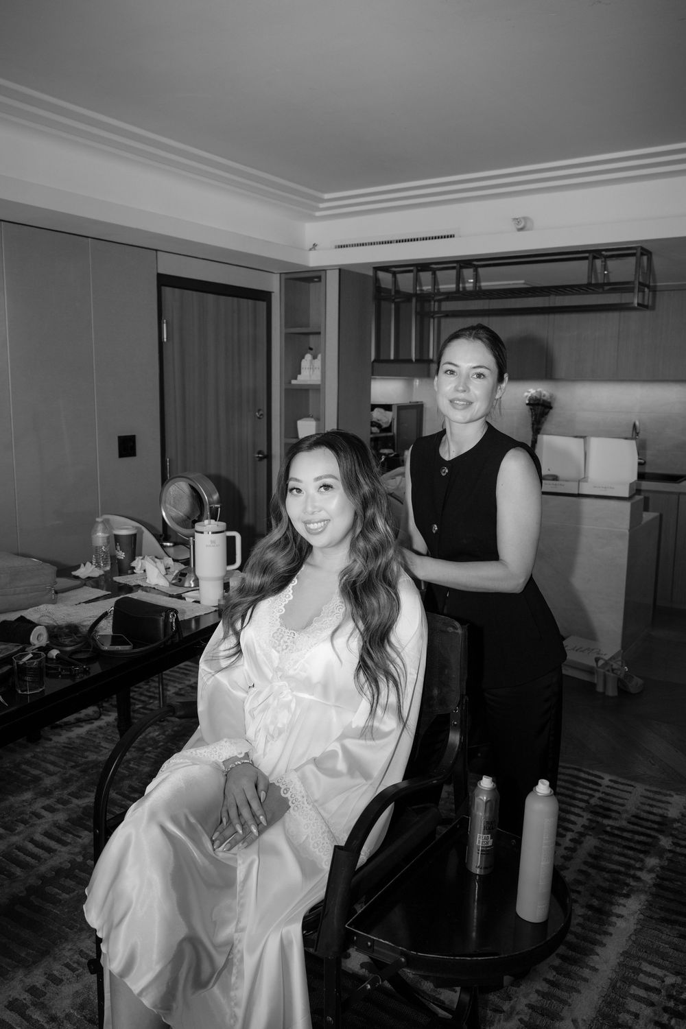 Black and white photo of a bride in a satin robe getting her hair styled by a beautician in a hotel room.