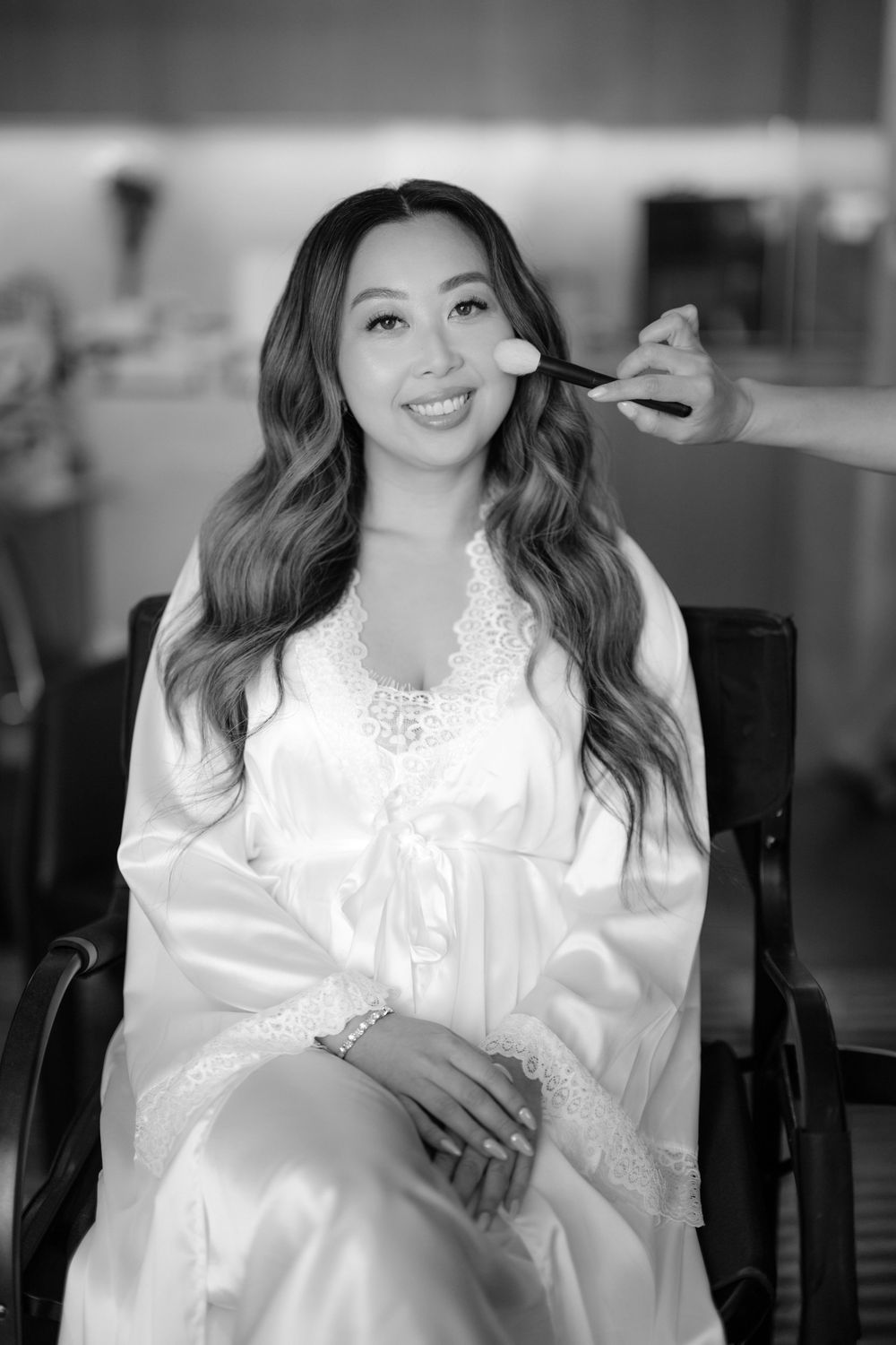 Black and white photo of a smiling bride in a satin robe getting makeup applied with a brush.