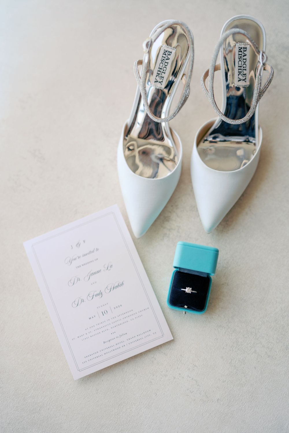 White Badgley Mischka bridal heels beside a wedding invitation and teal ring box with diamond engagement ring.
