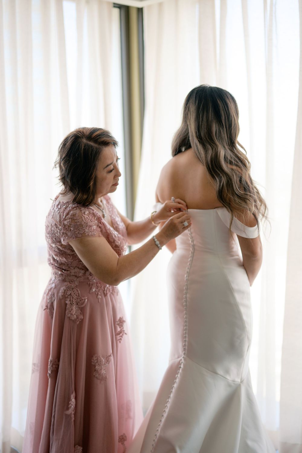 Mother helping bride button her white off-shoulder wedding gown in front of sheer curtained window.
