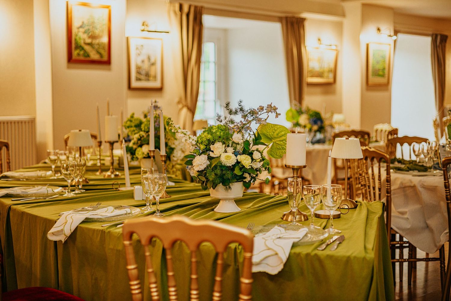 Elegant green tablecloth wedding reception table with white floral centerpiece, crystal glasses, and gold chairs in a warmly lit venue.