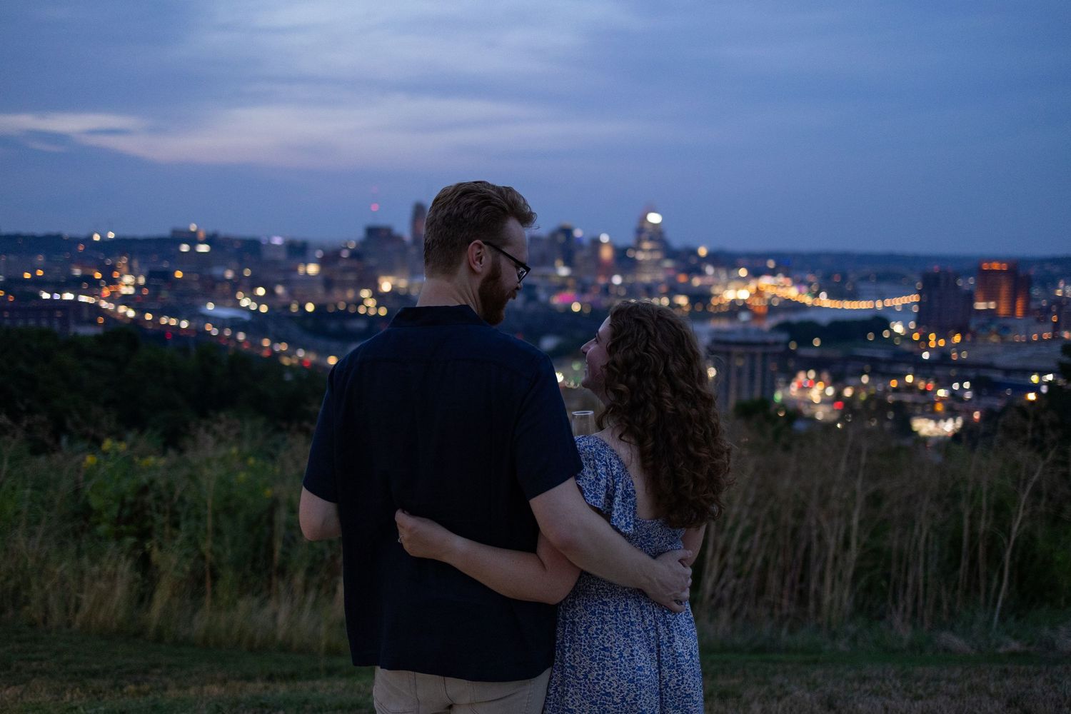 Couple embracing at dusk overlooking a glowing city skyline from a grassy hilltop.