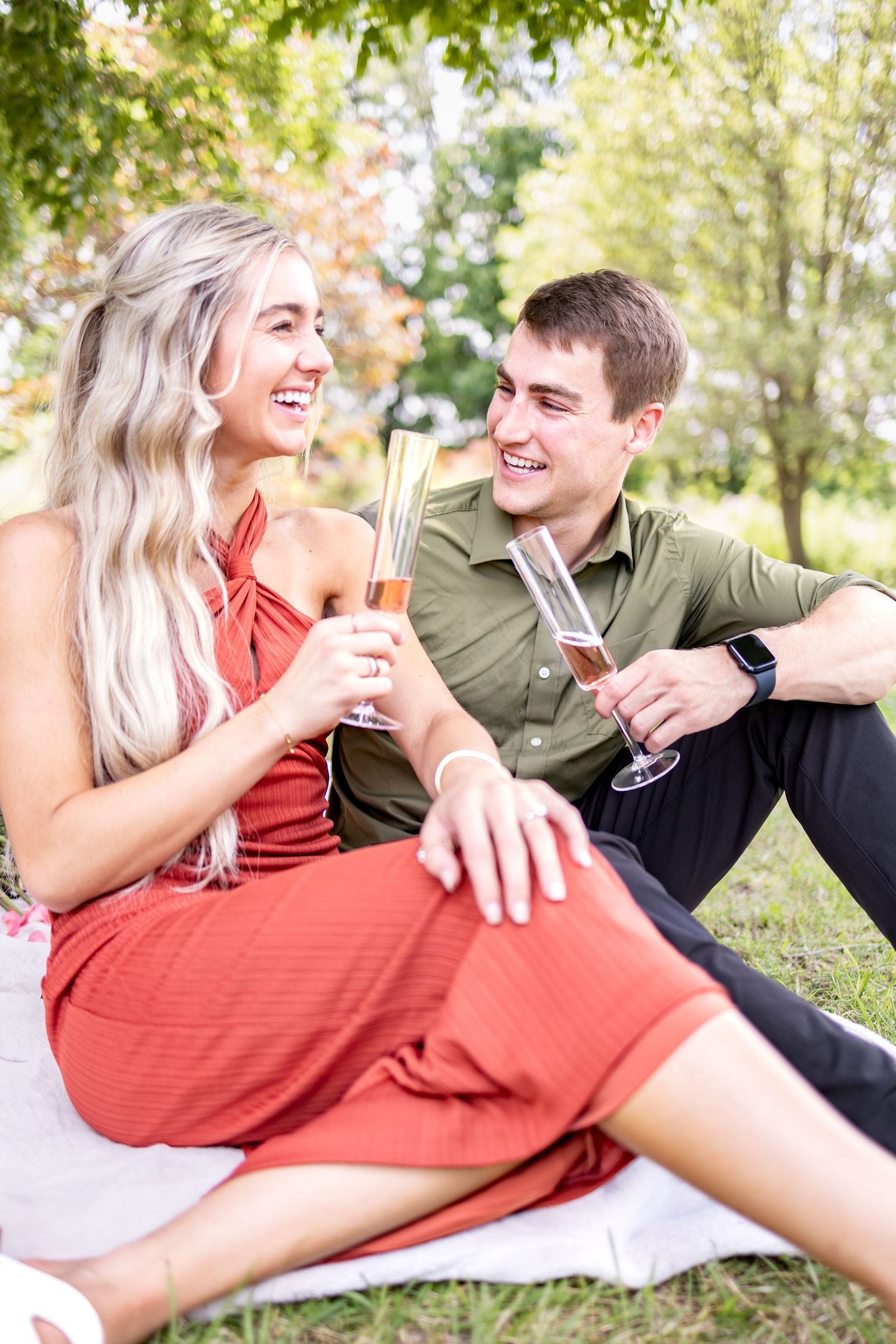 A smiling couple celebrates with champagne on a picnic blanket in a lush green Voice of America Park, dressed in red and olive outfits.