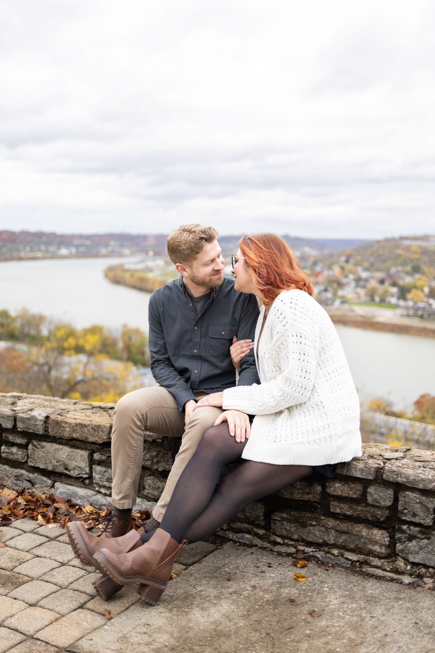 Couple sitting on stone wall overlooking the Ohio river valley in autumn, sharing a romantic moment at Twin Lakes in Eden Park Cincinnati OH