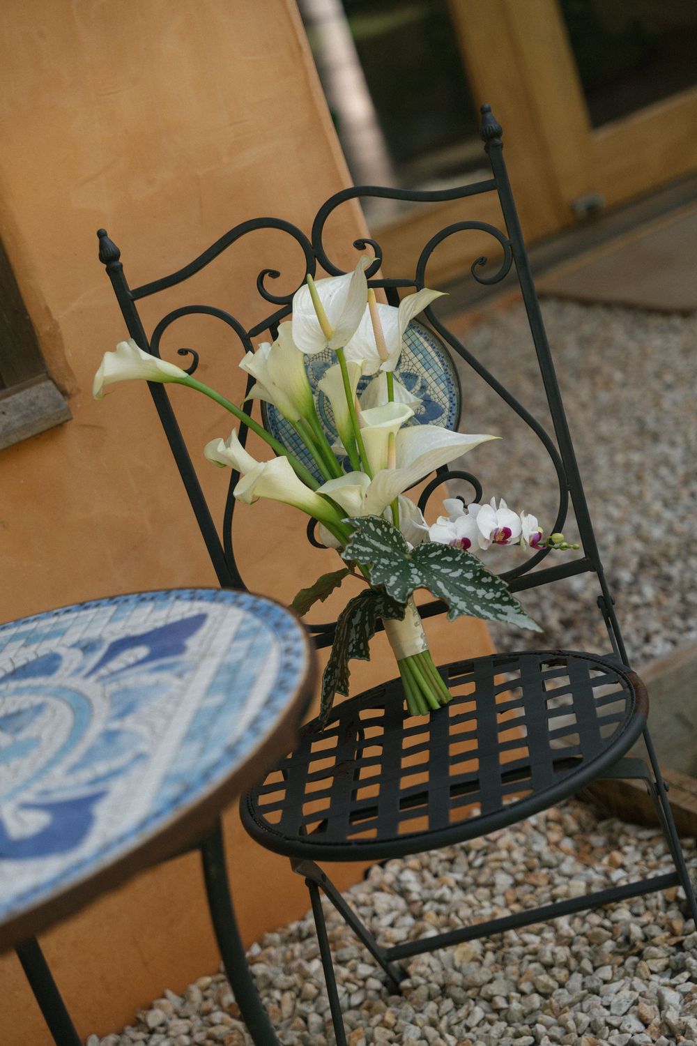 White calla lily wedding bouquet resting on a black wrought iron chair beside a blue mosaic table on a gravel patio.