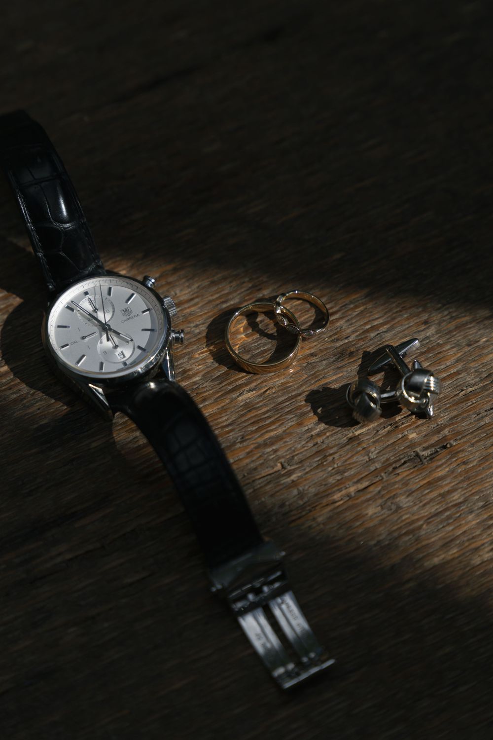 A luxury chronograph watch with black leather strap beside gold rings and silver cufflinks on a wooden surface.