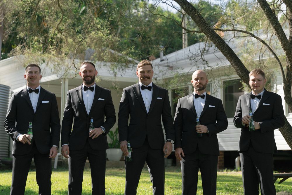 Five groomsmen in black tuxedos with bow ties smiling outdoors at a wedding venue surrounded by trees.