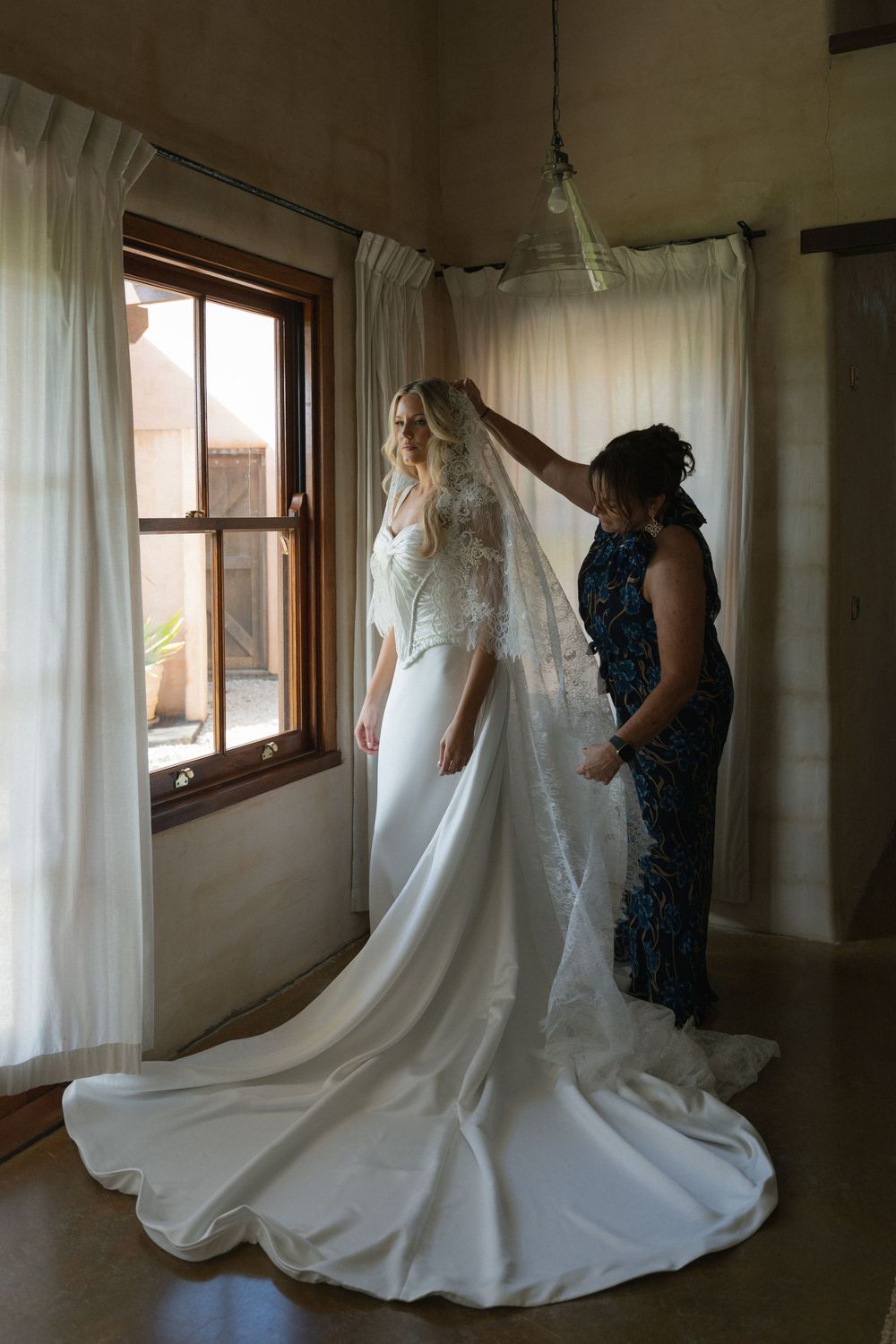 A bride in a white gown and lace veil being helped by a woman in a blue floral dress near a sunlit window.