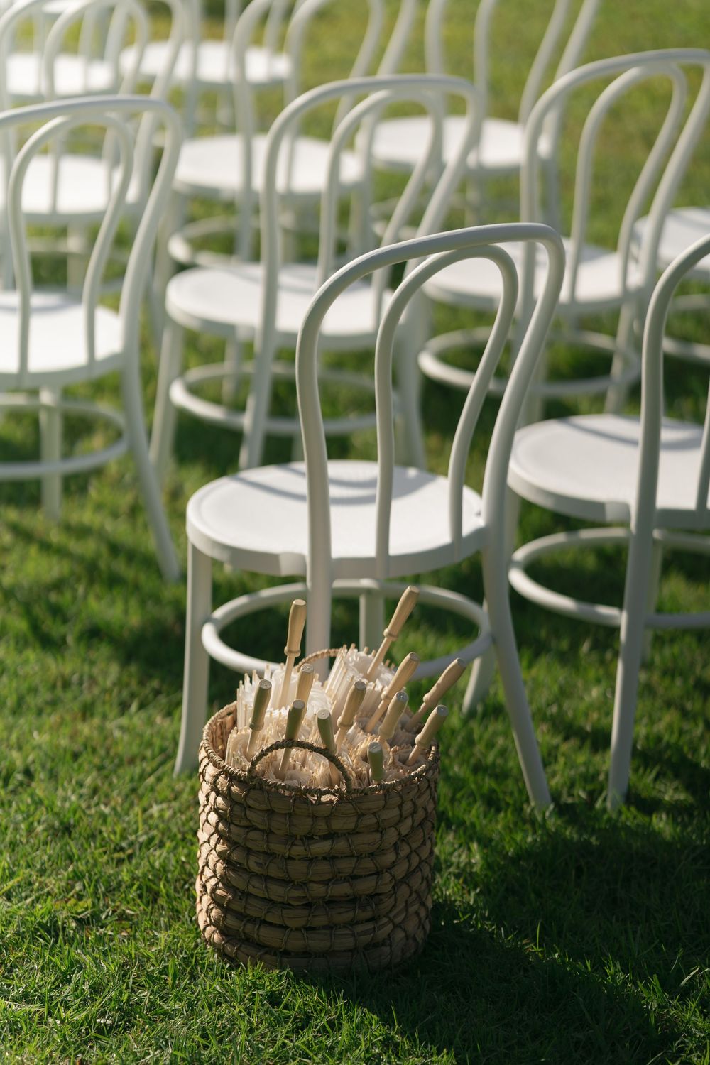 White bentwood chairs arranged on green grass with a wicker basket of hand fans for an outdoor wedding ceremony.