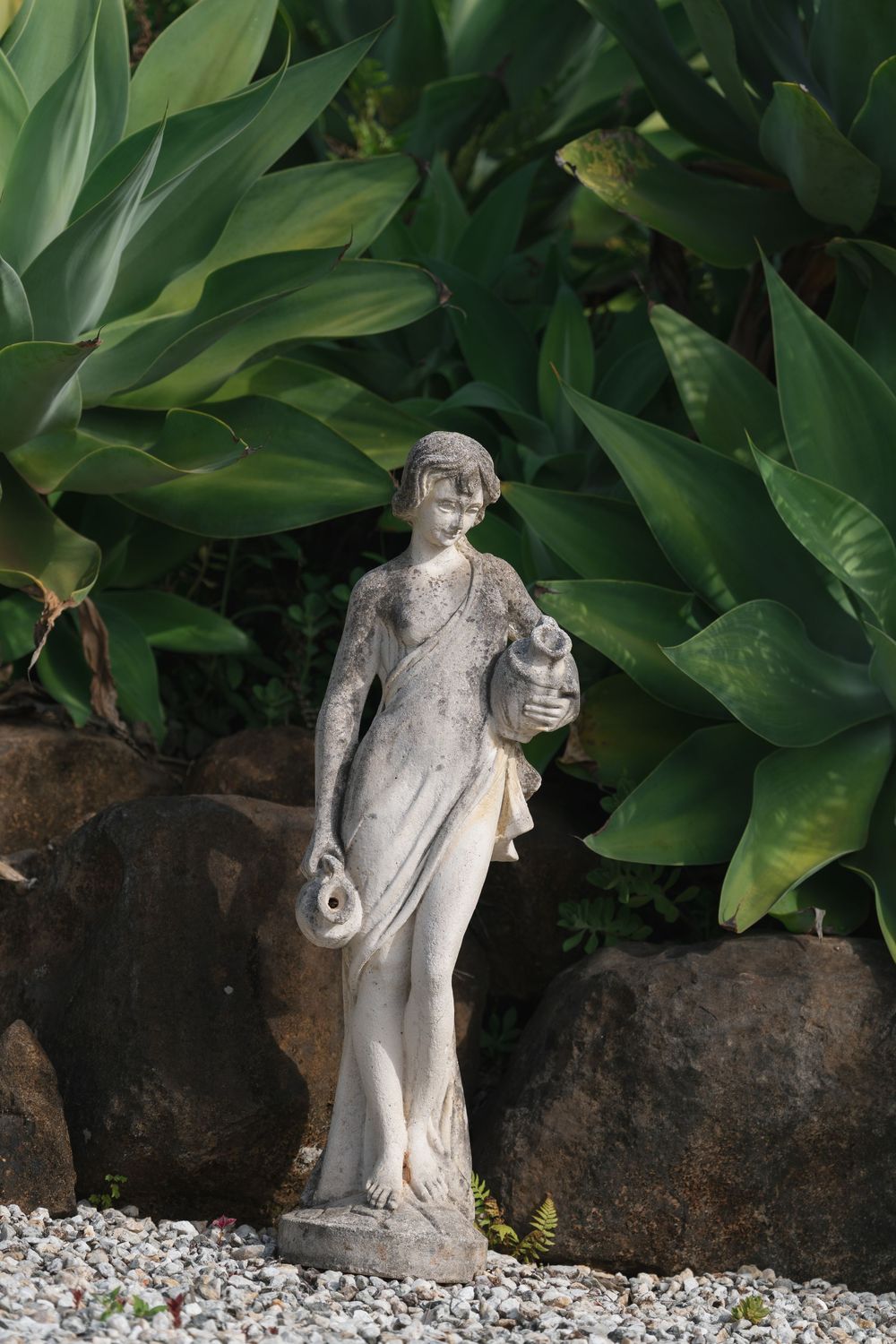 A weathered stone garden statue of a woman holding a vase, surrounded by lush tropical green leaves and rocks.