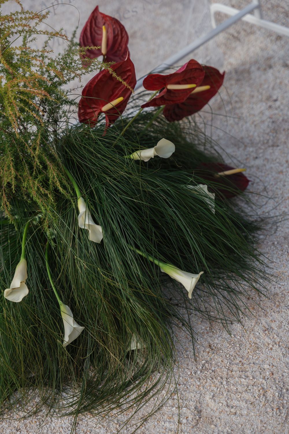 Elegant wedding bouquet with white calla lilies, red anthuriums, and lush green grass arranged on stone.
