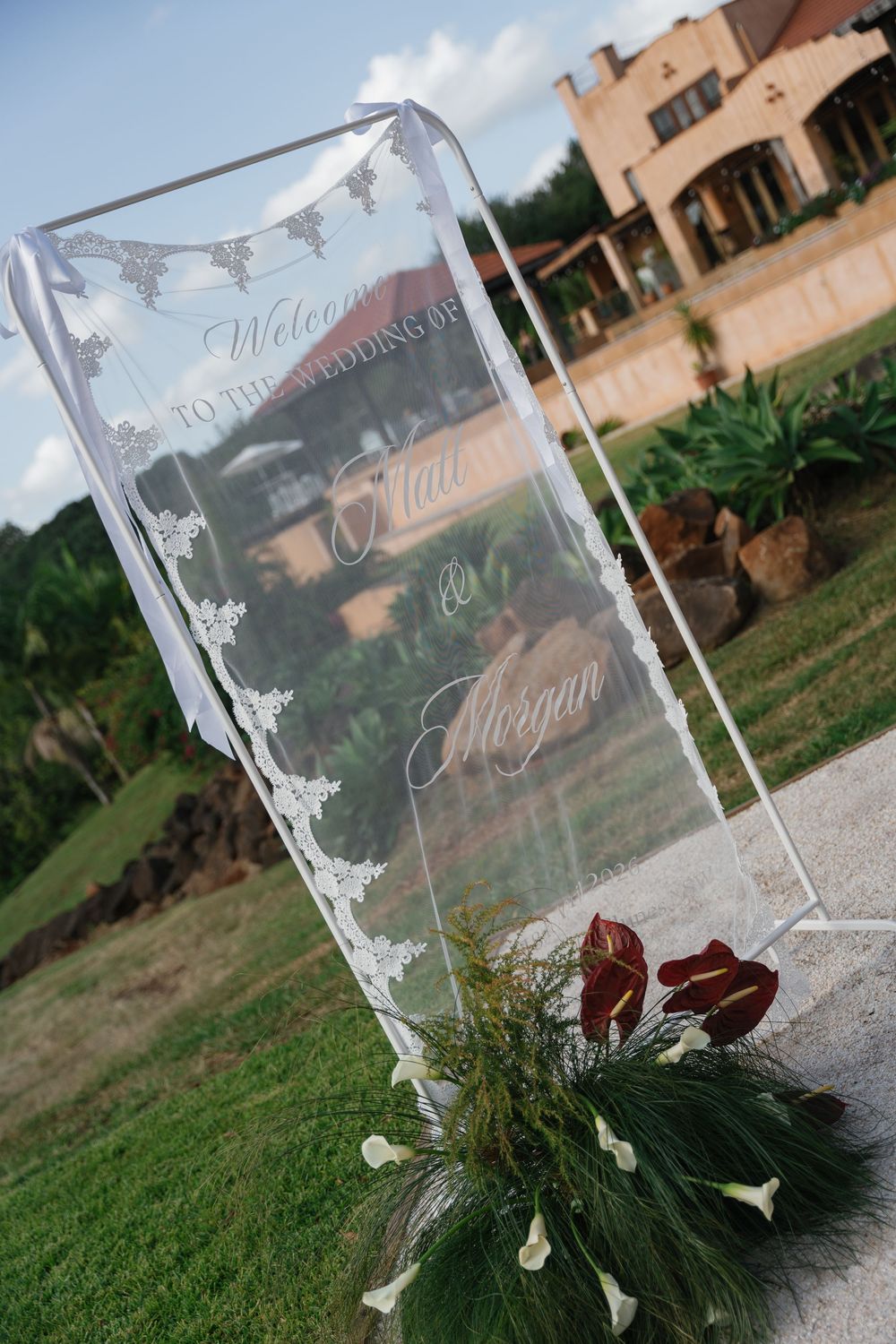 Elegant acrylic wedding welcome sign reading Welcome to the Wedding of Marcus and Morgan with floral arrangements outdoors.