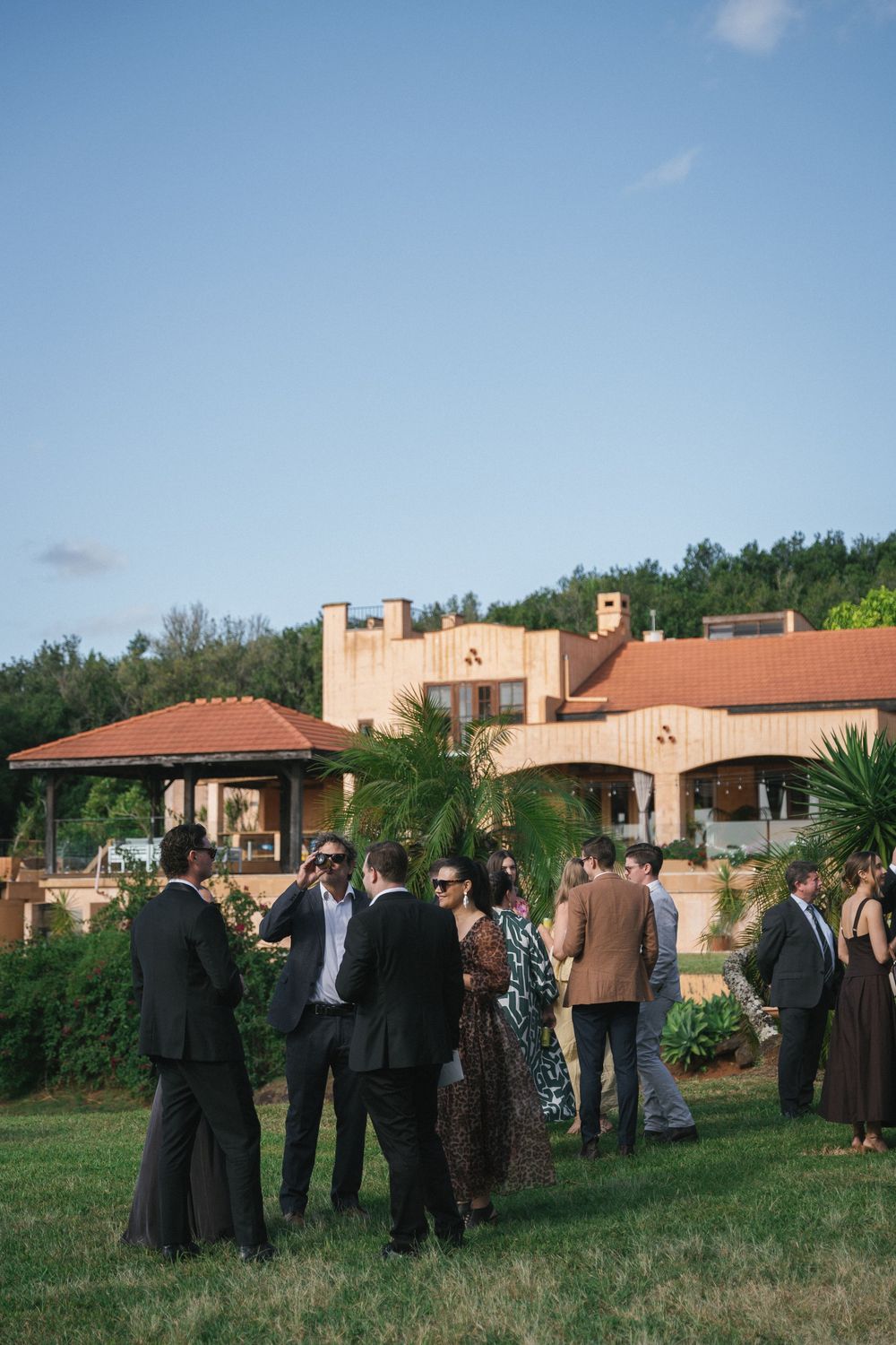 Elegantly dressed wedding guests mingle on green lawn outside rustic villa with terracotta roof and lush hillside backdrop.