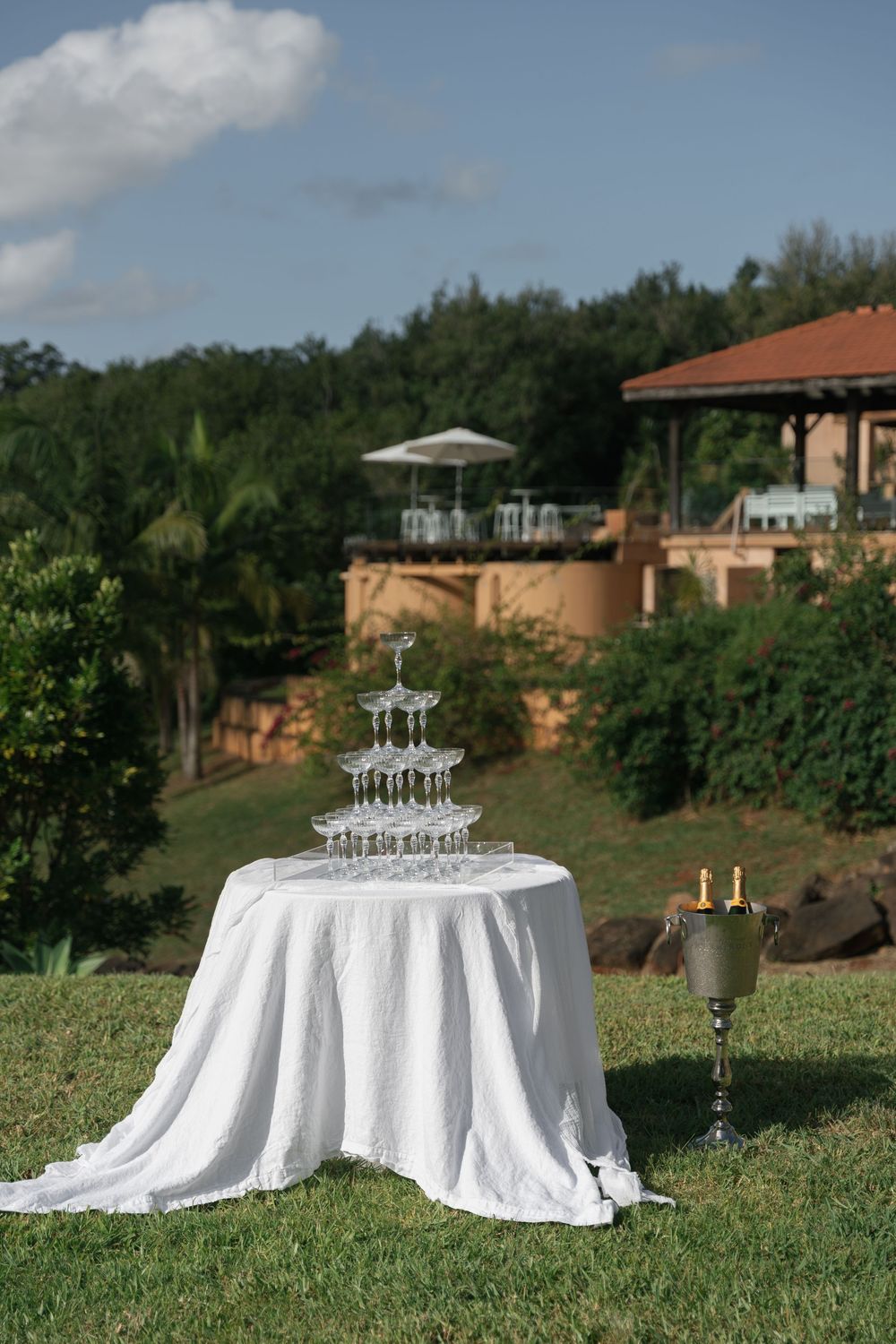 Champagne tower on white linen table with candelabra on manicured lawn at outdoor wedding venue with wooded hills.