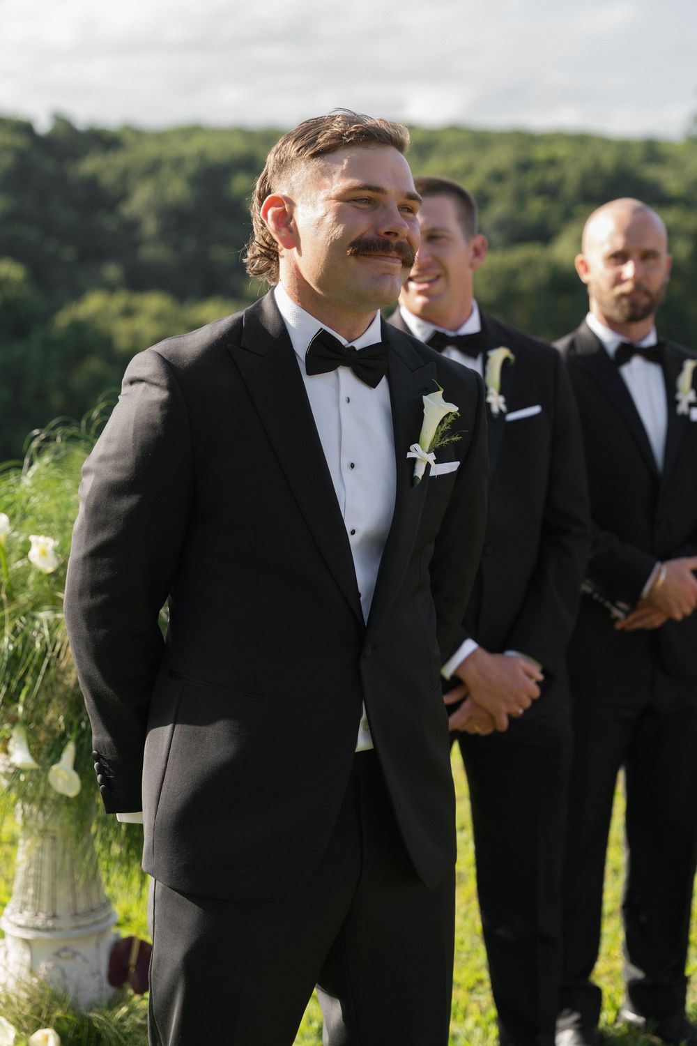 Groom in black tuxedo with bow tie smiling at altar with groomsmen standing behind him outdoors.