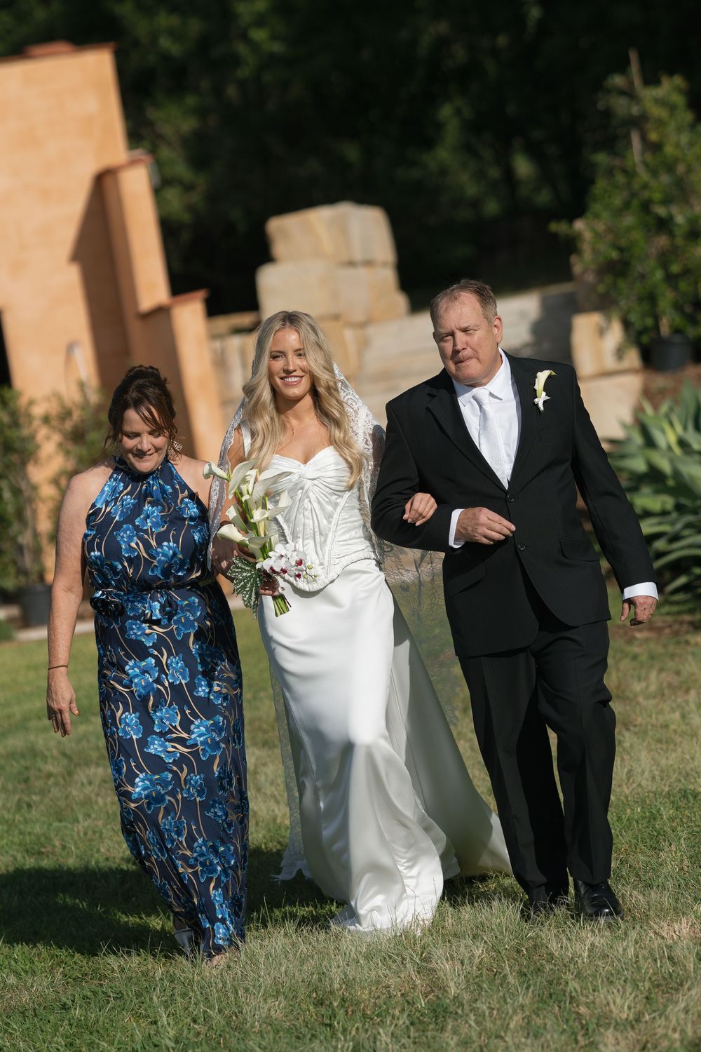 Bride in white gown walks down aisle with parents at outdoor wedding ceremony on sunny day.