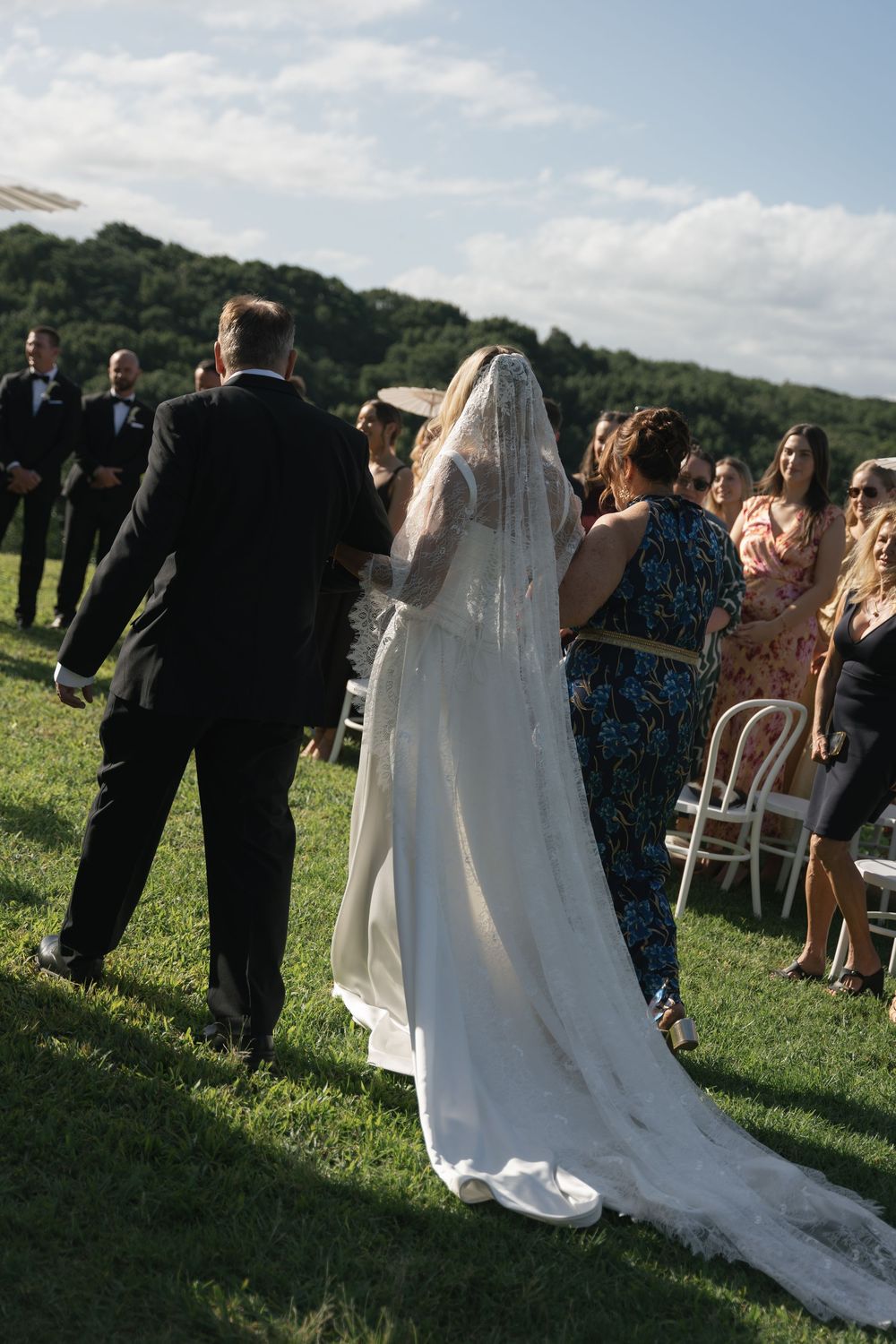 Bride in white gown and veil walks down the aisle escorted by two people at an outdoor wedding ceremony.