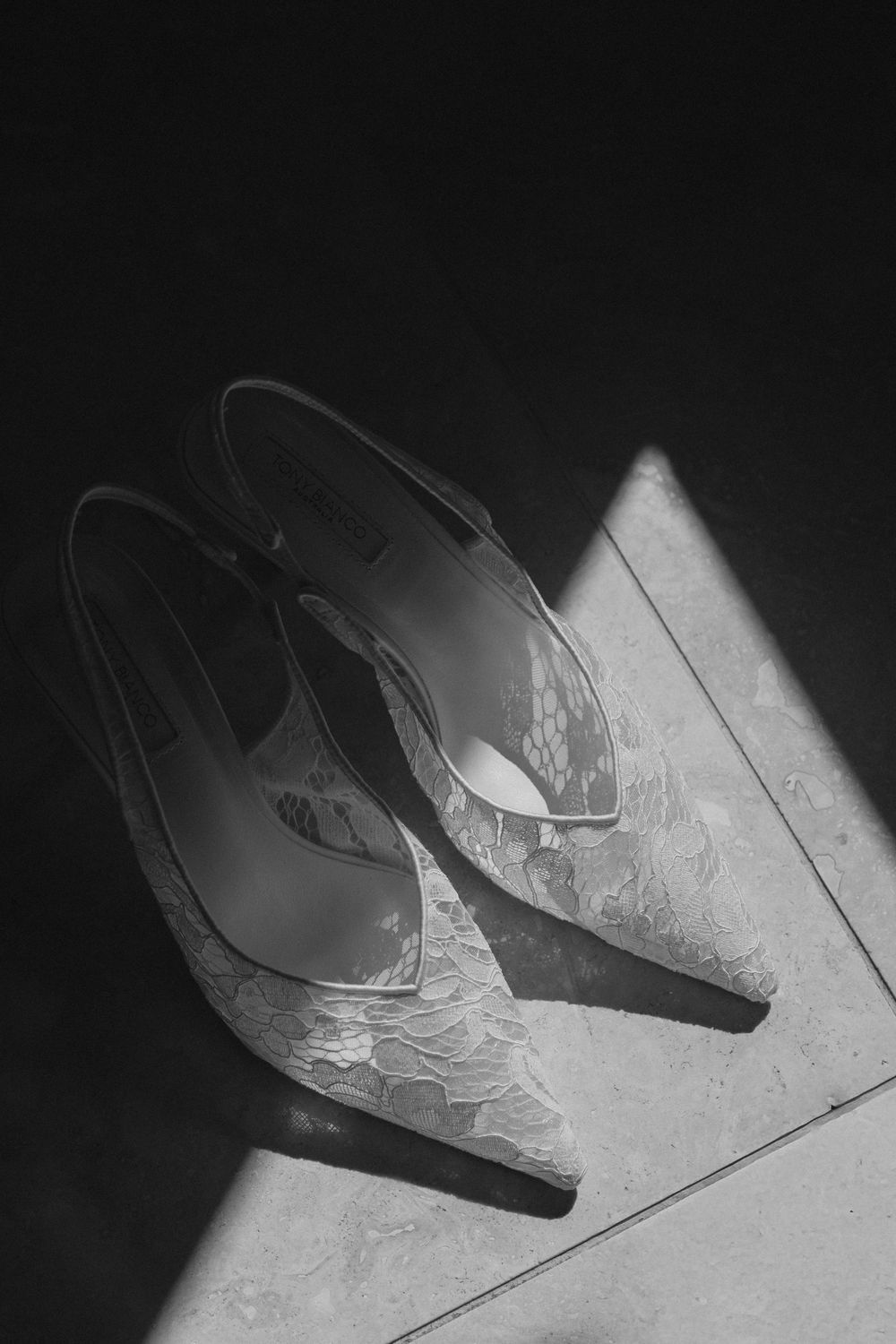 Black and white photo of elegant lace bridal heels resting on a tiled floor in dramatic natural light.