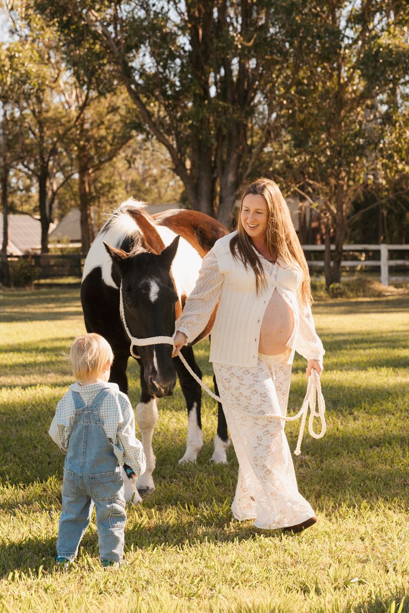 Pregnant woman in white outfit leads paint horse on a sunny farm while a toddler in denim overalls stands nearby during a family photoshoot