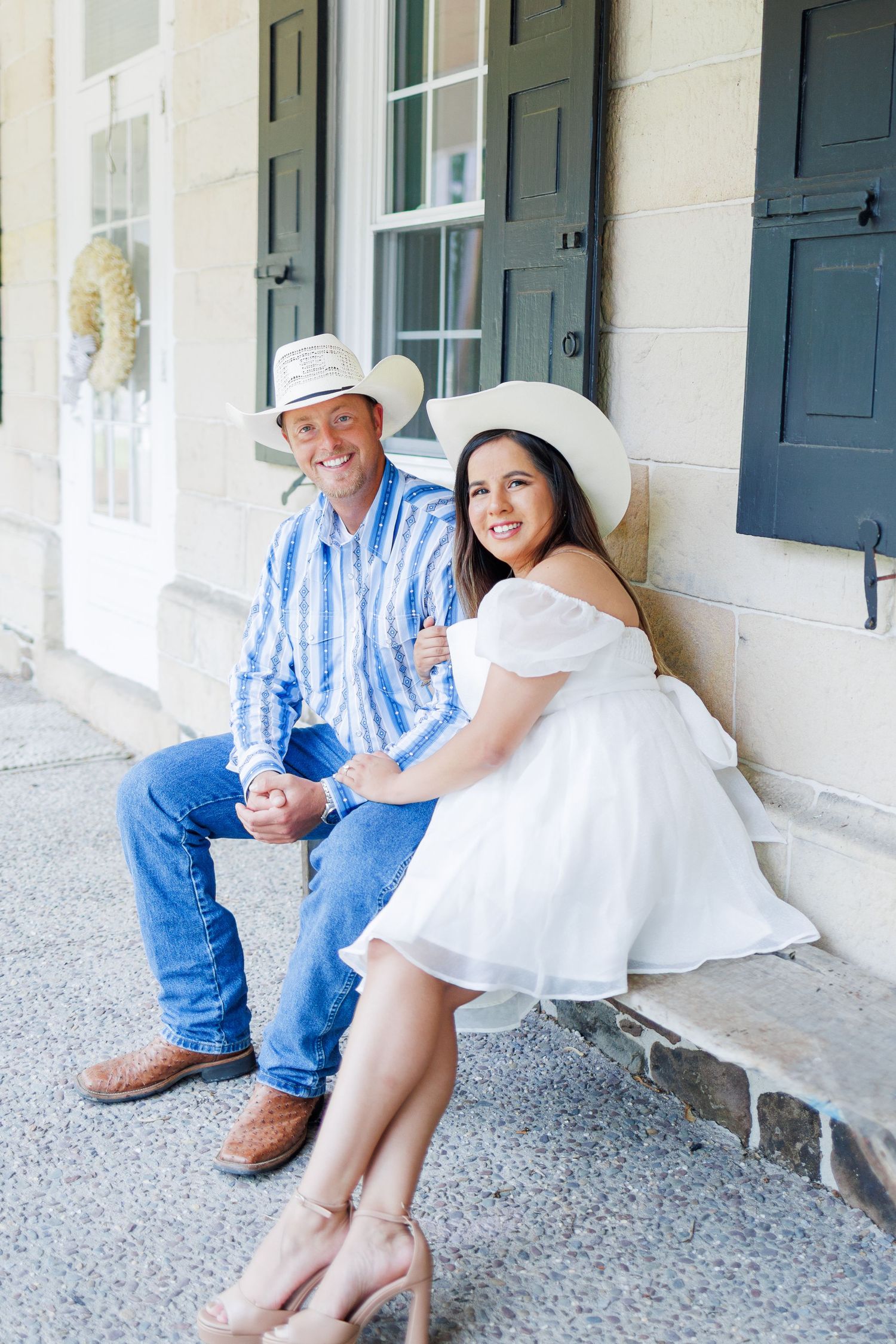 A smiling couple in cowboy hats sit together on steps, man in blue shirt and jeans, woman in white dress.