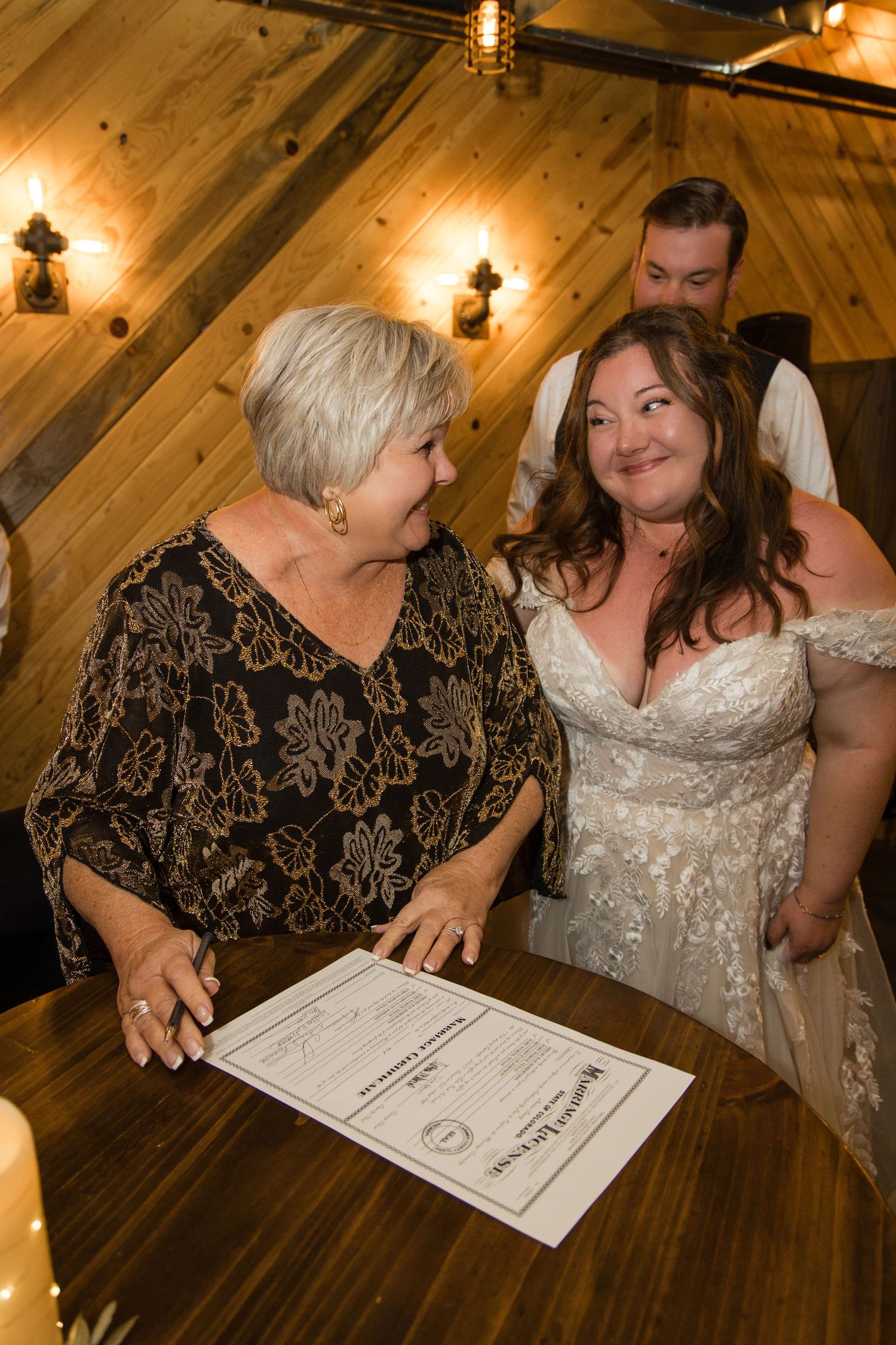 Bride in lace gown smiling at older woman signing marriage certificate at wooden table in rustic venue.