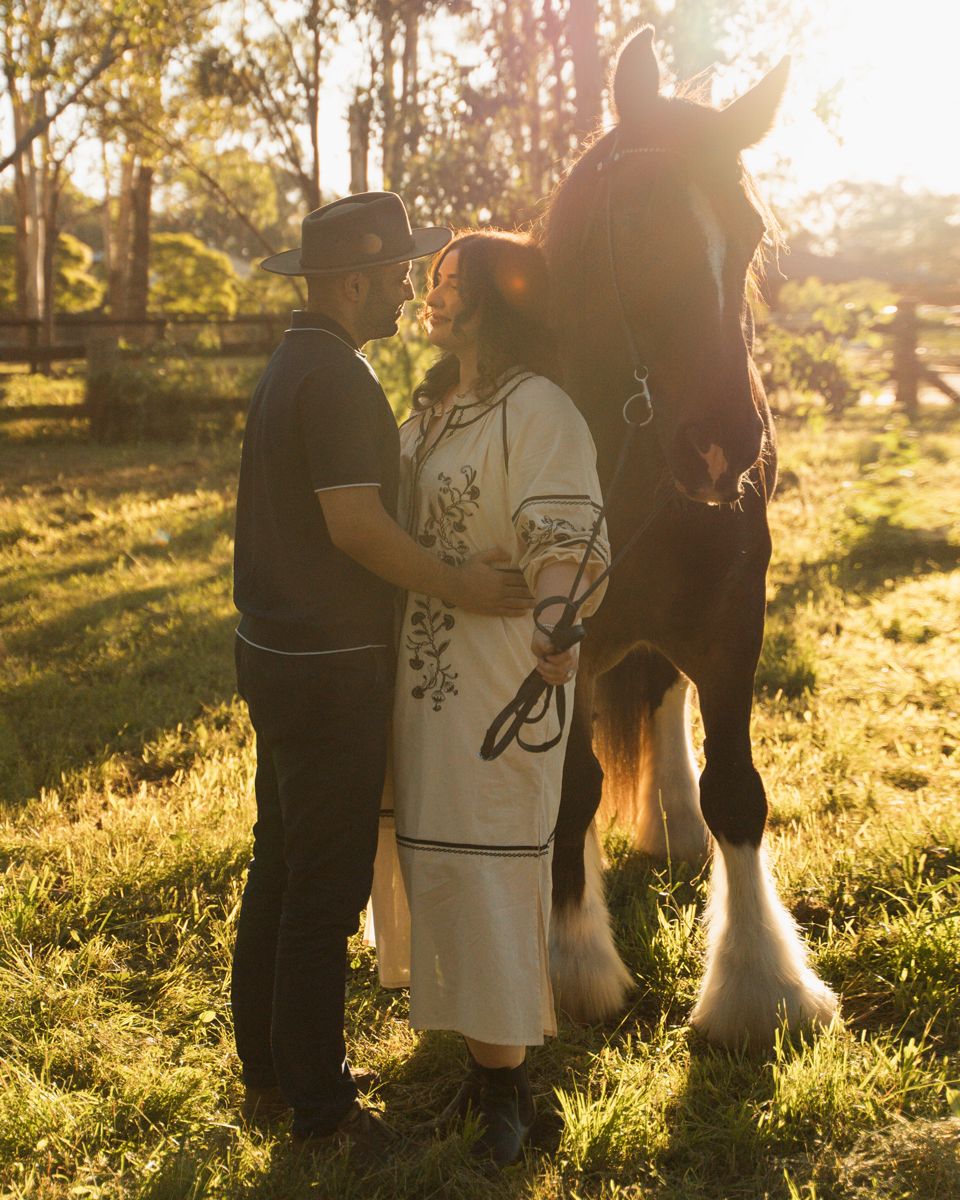 Couple standing with a shire horse in a sunlit green pasture, woman in white dress, man in black polo. Photoshoot with a horse in Sydney
