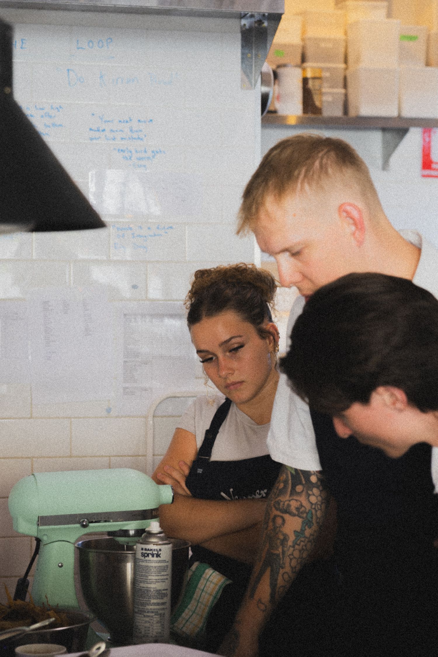 Three kitchen workers in aprons collaborate around a mint green stand mixer in a busy restaurant kitchen.
