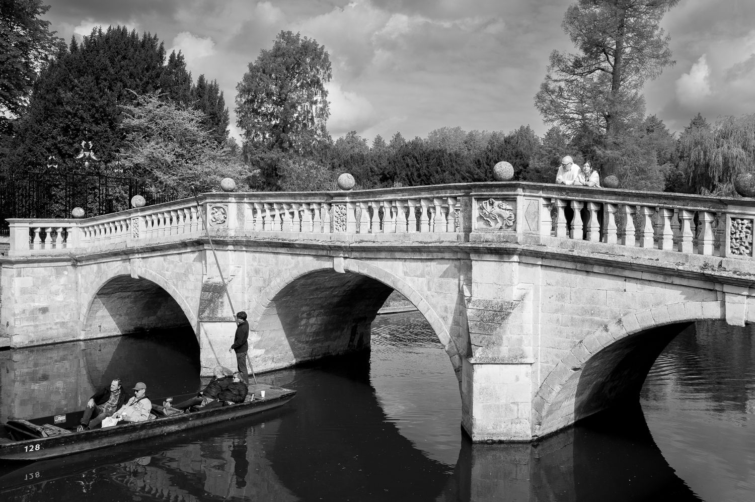 Black and white photo of people punting under a elegant stone bridge with arched spans in a serene park waterway.