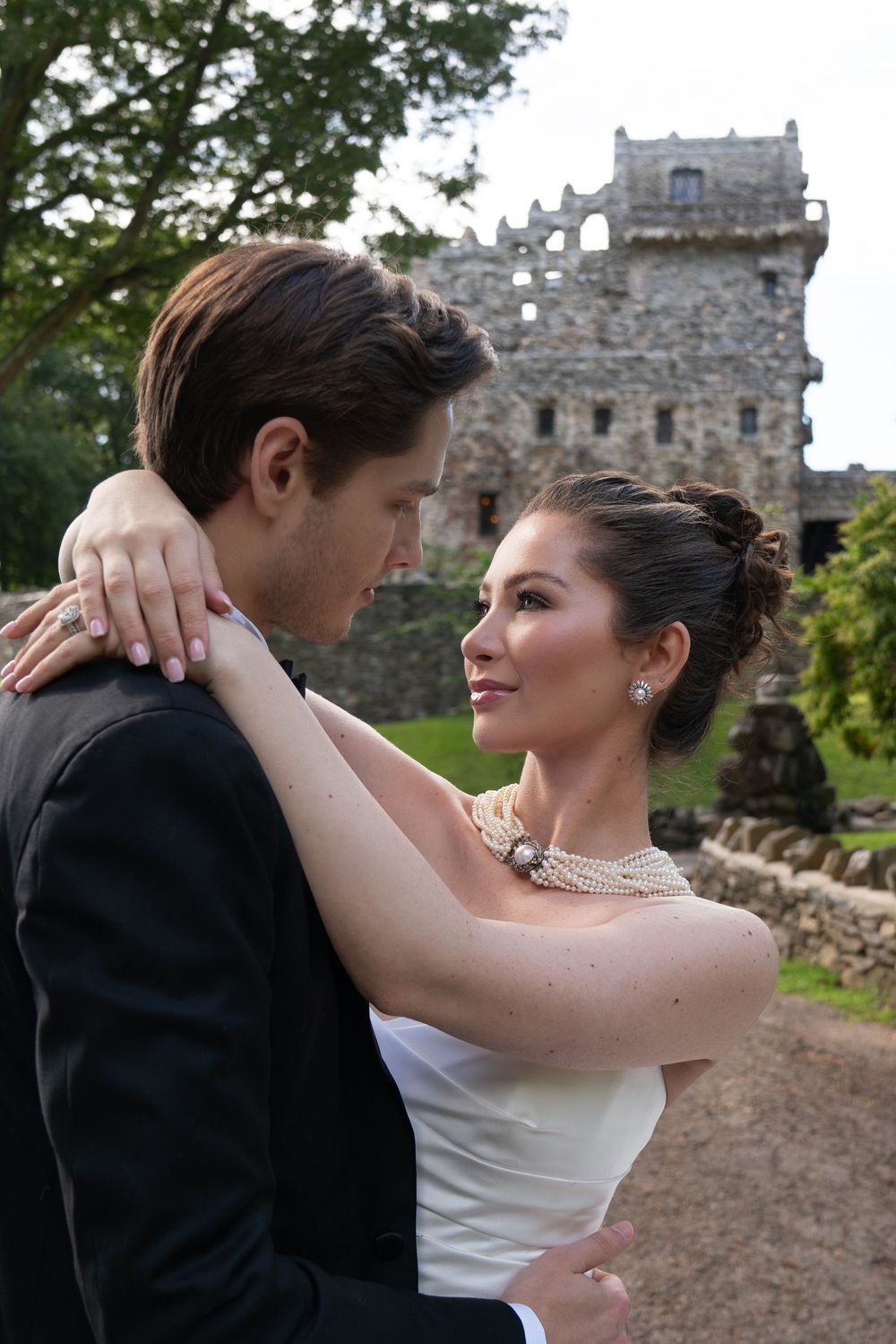 Bride in strapless gown with pearl necklace embraces groom before medieval stone castle ruins.