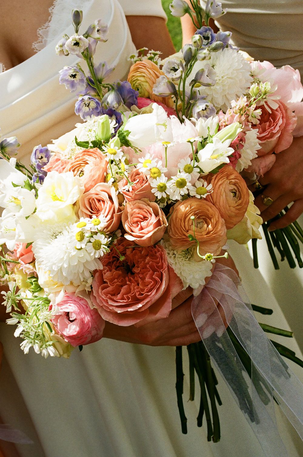 Close-up of bridal bouquet with coral roses, ranunculus, daisies, and blue wildflowers.
