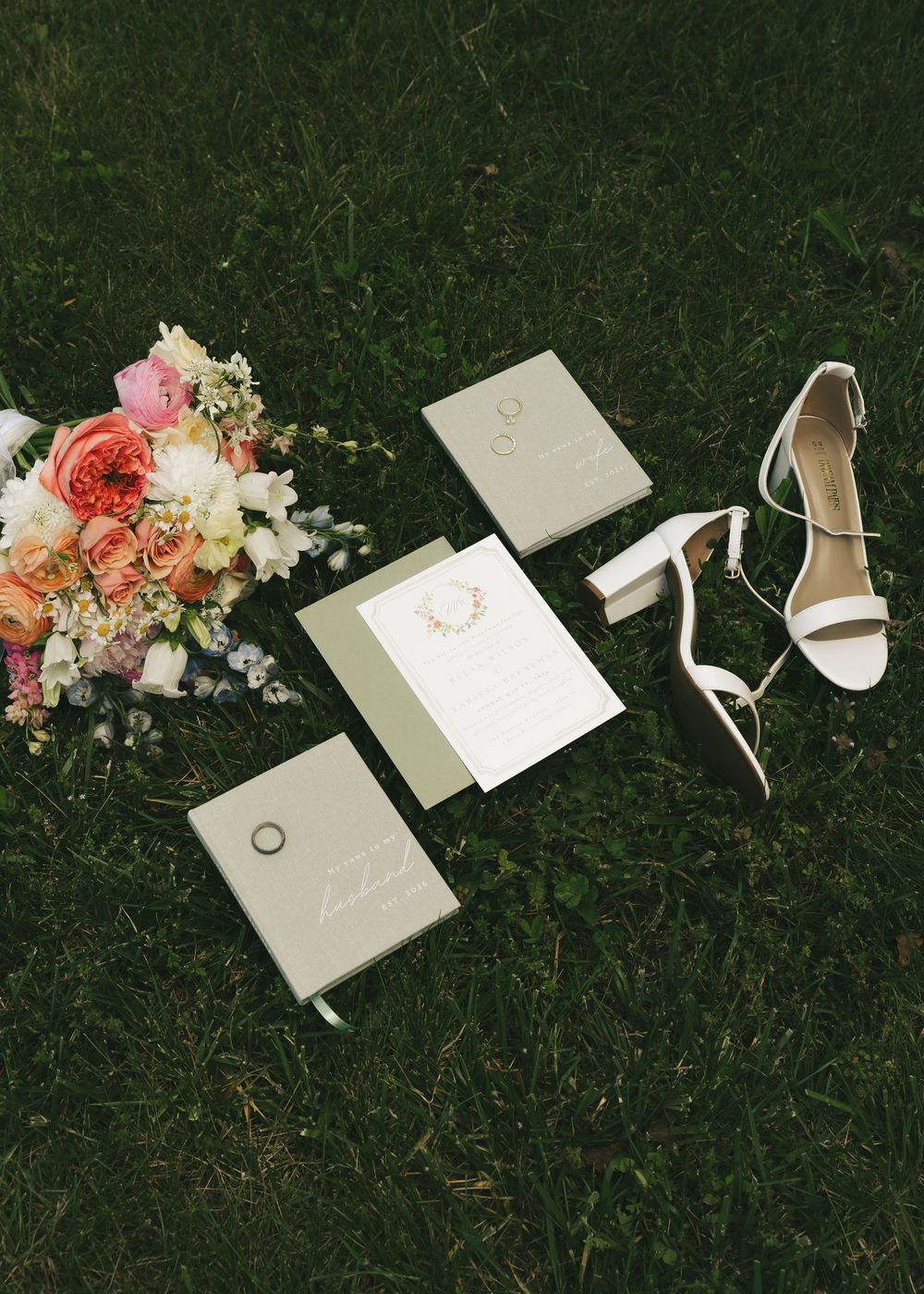 Wedding flat lay on grass featuring bridal heels, sage green stationery, ring box, and a colorful peach floral bouquet.