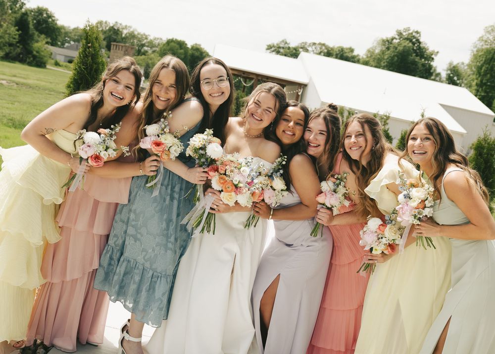 Group of eight bridesmaids in mismatched pastel dresses smile and hold bouquets together at an outdoor wedding venue.