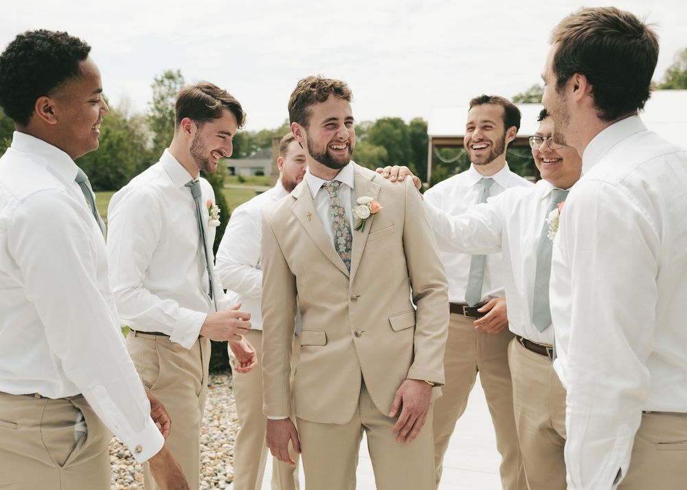 Groomsmen in white shirts and khaki pants laughing with groom in tan suit at outdoor wedding.