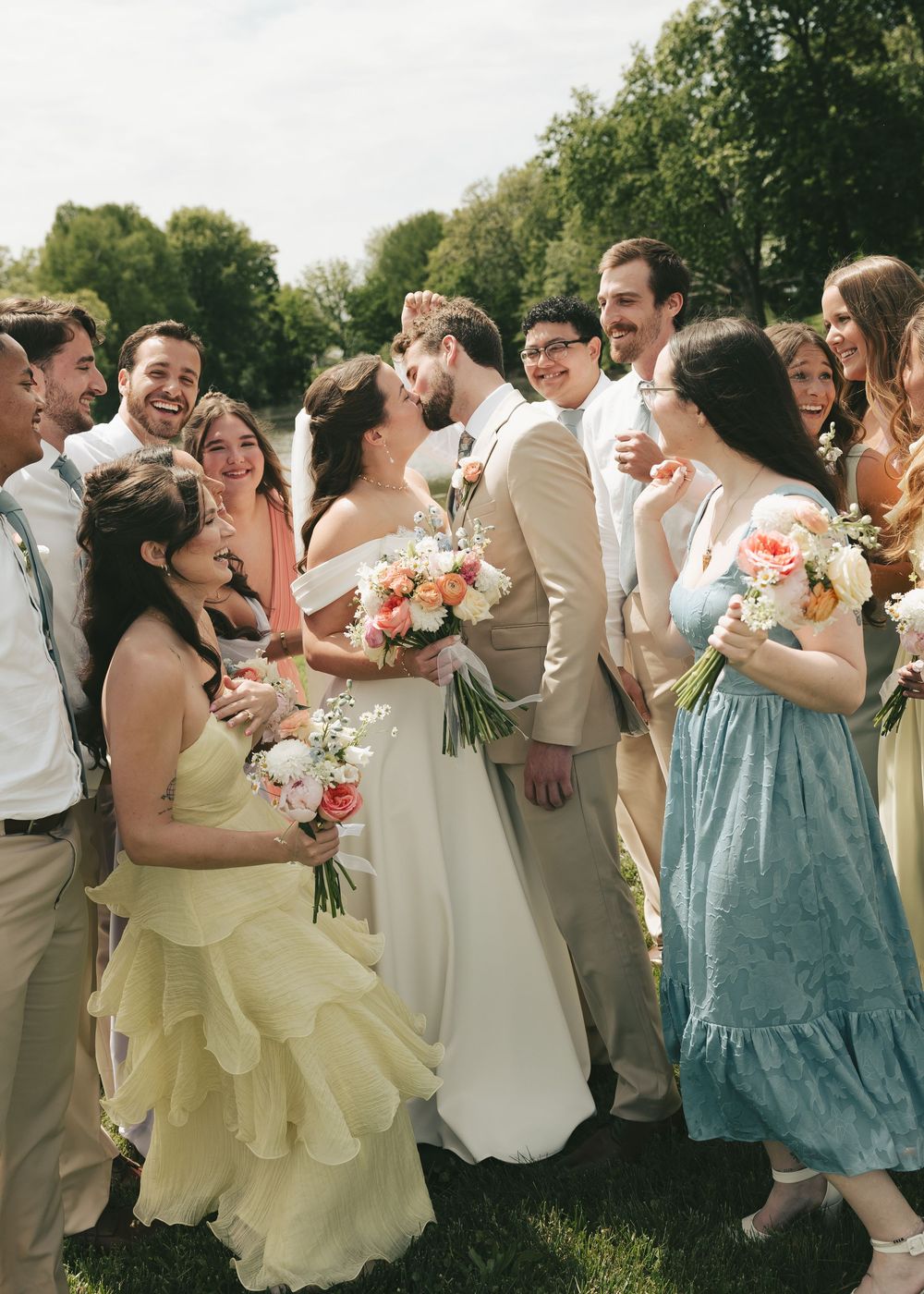 Wedding party surrounds kissing bride and groom outdoors near a lake on a sunny day.