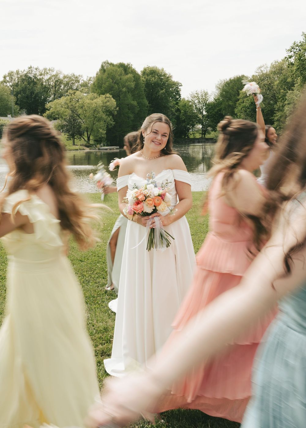 Bride in off-shoulder white gown holds colorful bouquet surrounded by bridesmaids in pastel dresses.