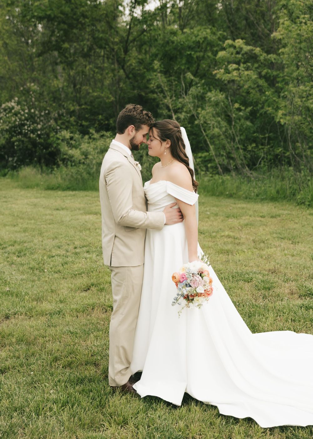 Bride in off-shoulder gown and groom in tan suit embrace in lush green meadow.