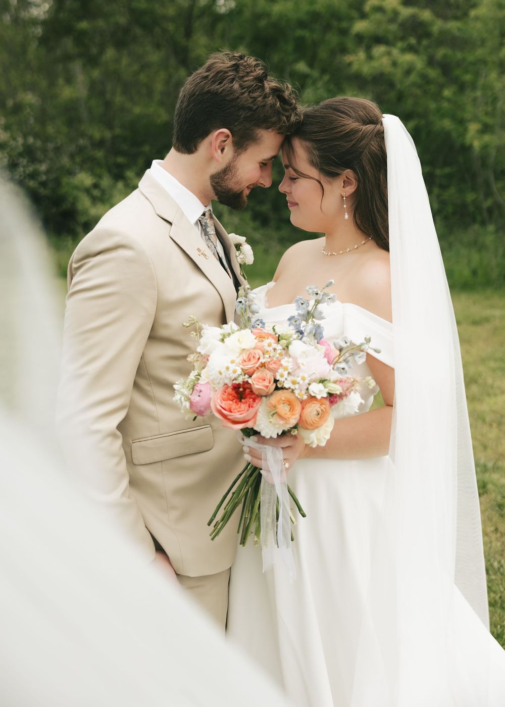Bride in white veil and groom in beige suit touch foreheads tenderly in outdoor portrait.