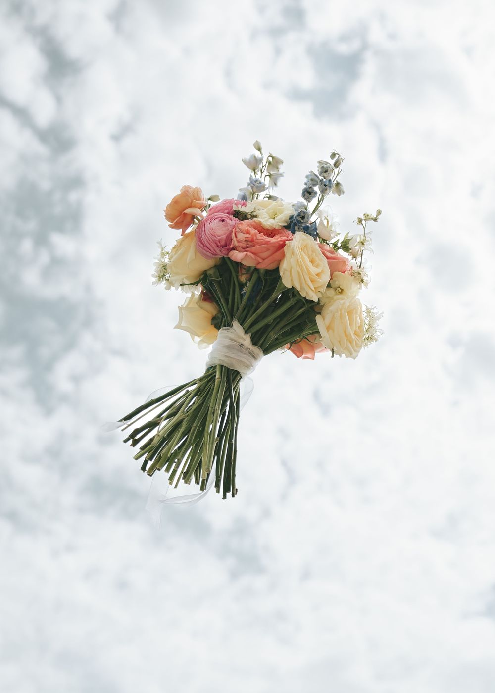 Wedding bouquet of coral, cream, and blue flowers tossed against a cloudy sky.