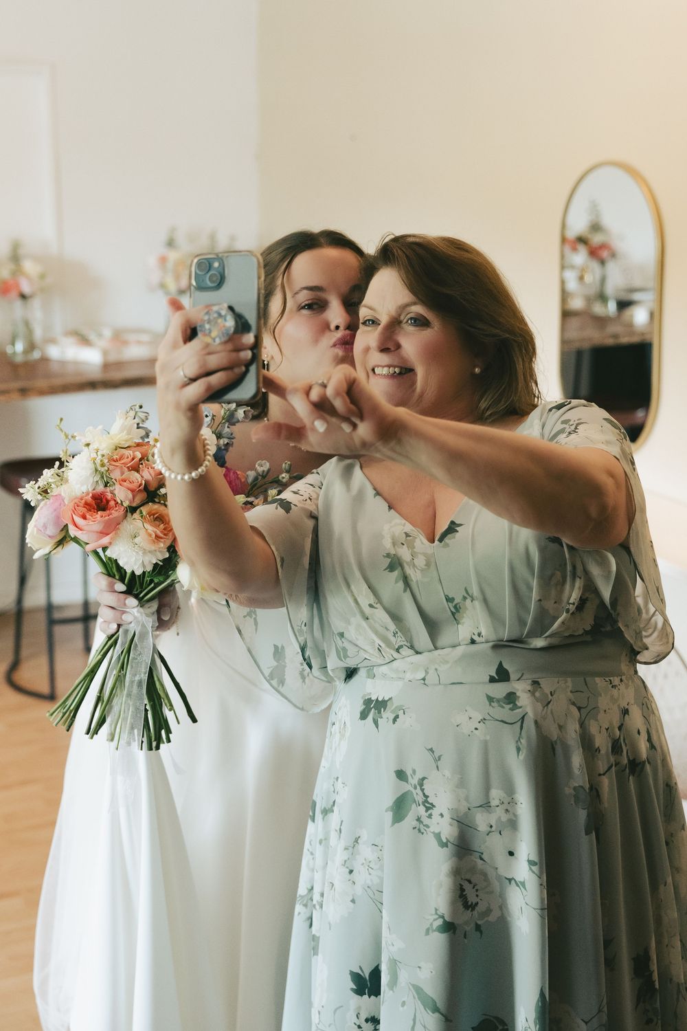 Bride in white dress and woman in floral gown take a selfie together while bride holds a pastel wedding bouquet.